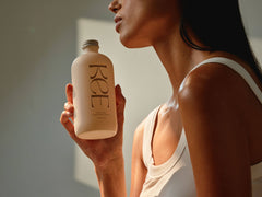 A woman in a white tank top holds a KEE Liquid Marine Collagen - Limoncello bottle, highlighting its benefits.