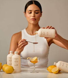 Woman pours KEE Liquid Marine Collagen - Limoncello onto a spoon above a glass with lemon slices; bottles and lemons nearby.