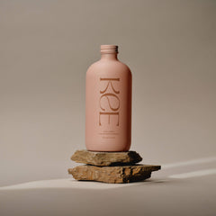 A pink KEE Liquid Marine Collagen - Mango Sorbet bottle stands on stacked rocks with a softly lit neutral background.