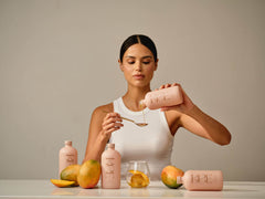A woman pours KEE Liquid Marine Collagen - Mango Sorbet into a spoon over a glass, with mangoes on the table.