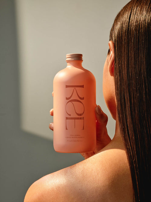 A person with straight brown hair holds a pink KEE Liquid Marine Collagen - Peach Elderflower bottle, facing away.
