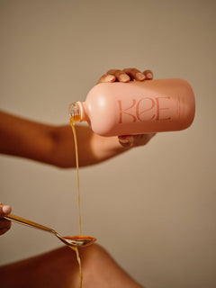 A hand pours KEE Liquid Marine Collagen - Peach Elderflower from a pink bottle into a gold spoon.