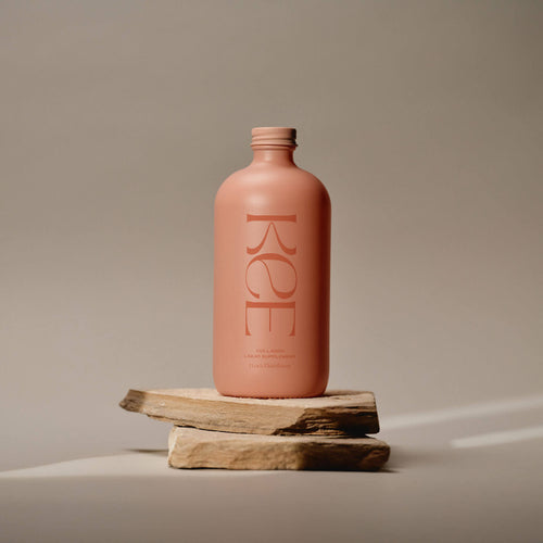 A matte pink bottle labeled KEE—Liquid Marine Collagen - Peach Elderflower—stands on stacked flat stones.