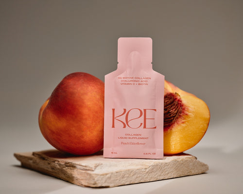 A KEE Liquid Marine Collagen Sachet - Peach Elderflower stands before a whole and halved peach on a stone surface.