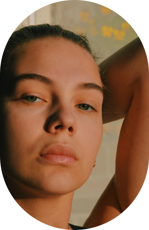 Close-up of a person with light eyes and natural makeup, gazing at the camera with an arm raised behind their head.