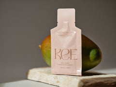 A KEE Liquid Marine Collagen Sachet - Mango Sorbet stands before a mango on stone with a neutral background.