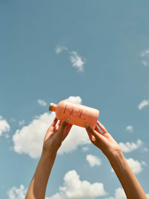 Two hands hold a pink bottle labeled KCEE up against a blue sky with scattered white clouds.