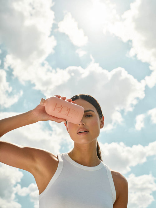 Woman in a white tank top holds a pink bottle labeled “KOE” against her forehead with a cloudy sky in the background.