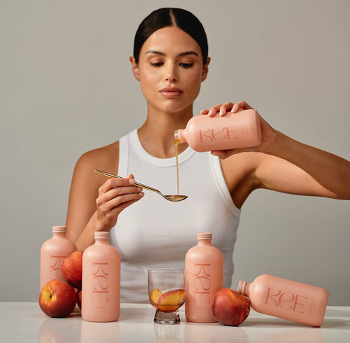 Woman holding a pink bottle and spooning a liquid into a glass with peaches around her.