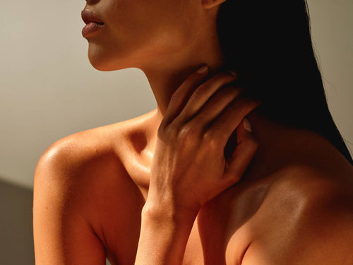 Close-up of a womans neck and shoulders with her hand gently touching her neck in warm, soft lighting.