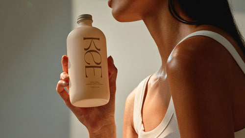 A woman in a white tank top holds a cream-colored bottle labeled Koe in soft natural light.